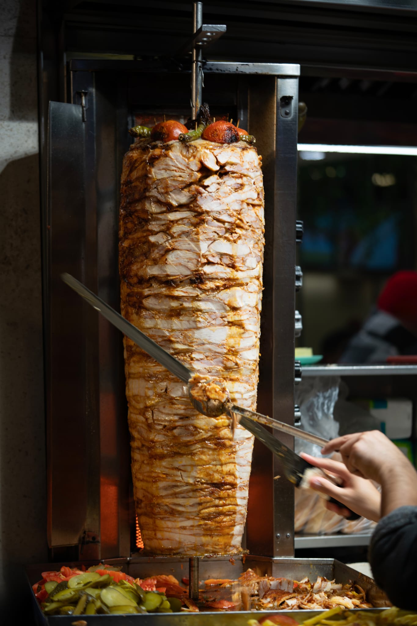Chicken Doner kebab being sliced on a vertical rotisserie with vegetables below.