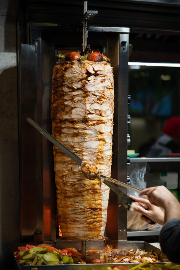 Chicken Doner kebab being sliced on a vertical rotisserie with vegetables below.